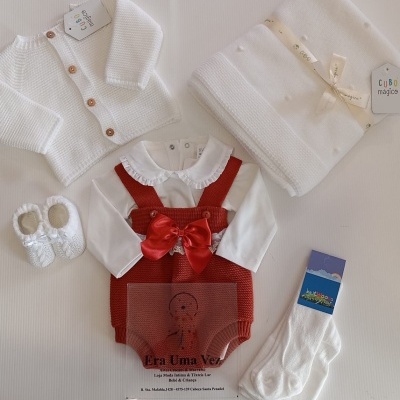 Conjunto de roupa de bebé com casaco branco malha, manta branca, sapatos renda, vestido com laço vermelho e camisa branca, meias brancas.