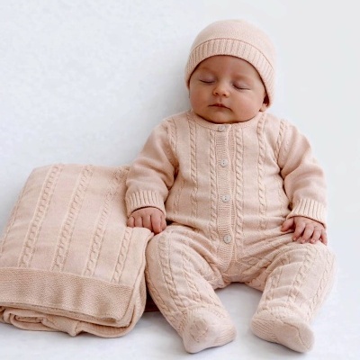 Bebé com fato e gorro de malha rosa claro sentado ao lado de manta rosa.