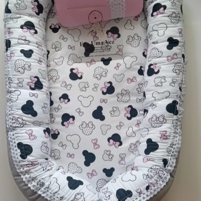 Ninho de bebê em tecido branco com estampas de cabeça de rato e laços cor-de-rosa, almofada rosa e detalhes em renda branca.