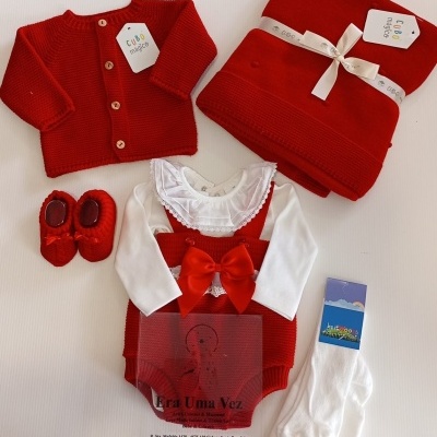 Conjunto de roupa de bebé num fundo branco com camisola vermelha, body branco, jardineira vermelha, manta, sapatinhos e meias.