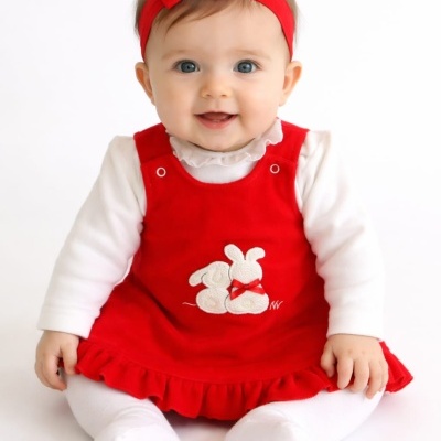 Bebé sorridente com vestido vermelho de coelho bordado, blusa e meias brancas, e faixa vermelha na cabeça
