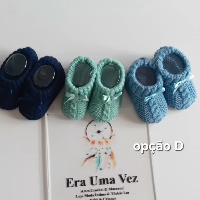 Três pares de sapatinhos de bebé em crochet em azul escuro, verde água e azul claro, com uma placa branca com texto e um apanhador de sonhos ao centro.