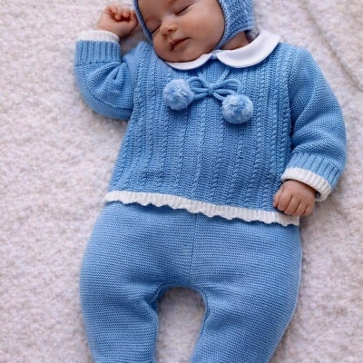 Bebé a dormir vestido com roupa de malha azul com detalhes brancos e chapéu a condizer, deitado numa manta felpuda branca.