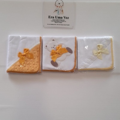 Três paninhos para bebé brancos com bordas e laços amarelos em embalagem transparente.