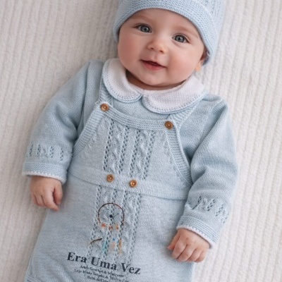 Bebé com macacão azul claro tricotado e gorro, deitado em superfície creme.