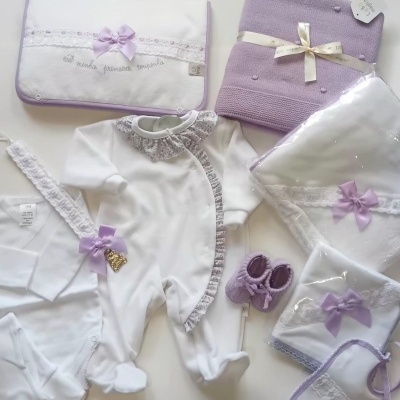 Conjunto para bebé branco e lilás com babygrow, manta, sapatinhos, fraldas e babete, com laços lilás e bordados.