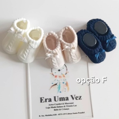 Três pares de sapatinhos de bebé em crochet branco, bege e azul escuro com laços sobre uma superfície branca.