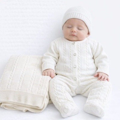 Bebé vestindo fato e gorro de malha branca sentado ao lado de manta branca dobrada