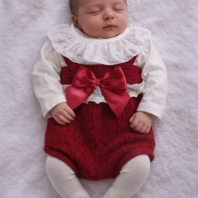 Bebé com roupa branca e jardineira vermelha com laço, deitado num fundo branco macio.