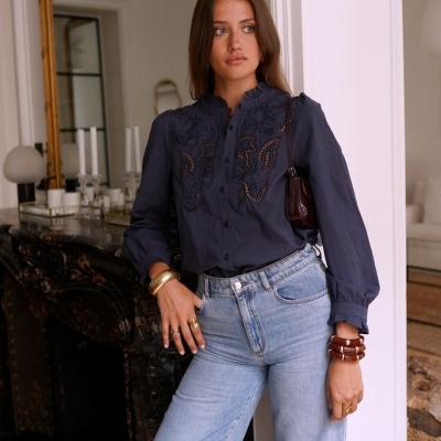 Mulher com blusa azul bordada e jeans azul claro, pulseiras e bolsa, em sala com espelho e lareira