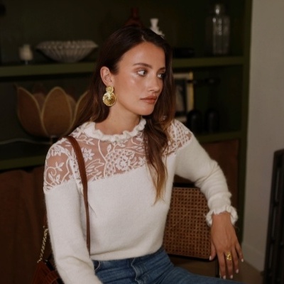 Mulher com camisola branca de renda e calças de ganga sentada em cadeira com parede e estante ao fundo