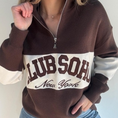 Camisola castanha e bege com fecho e texto preto, usada por pessoa com jeans azuis.