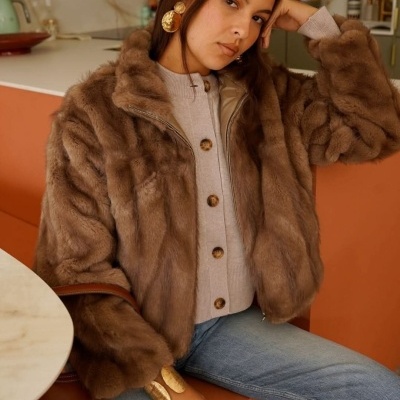 Mulher sentada com casaco felpudo castanho, cardigan bege e jeans azuis claros em cozinha