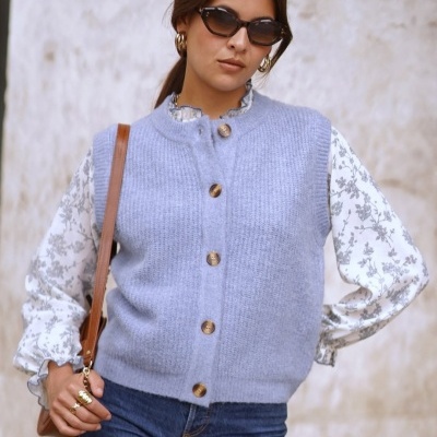 Mulher a usar colete cinza, blusa branca com padrão floral, jeans azuis, óculos escuros e mala castanha.