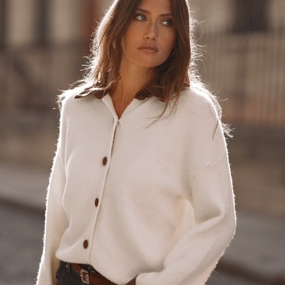 Suéter cardigan branco com botões castanhos, gola e punhos castanhos, jeans pretos e cinto castanho, em ambiente urbano.