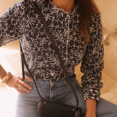 Mulher vestindo blusa preta floral branca, calças de ganga, bolsa preta com fivela dourada, e acessórios dourados.