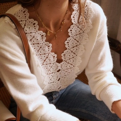 Mulher com camisola branca de malha com gola em renda, calças de ganga azuis e acessórios dourados sentada interior