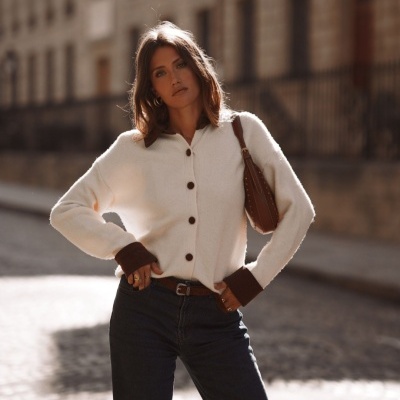 Mulher com camisola bege de malha com botões castanhos e punhos castanhos, calças de ganga escuras e mala castanha ao ombro na rua