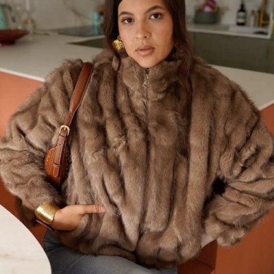 Mulher com casaco de pêlo castanho e acessórios dourados sentada numa cozinha moderna.