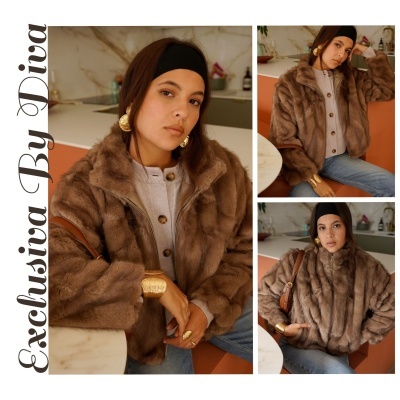 Mulher com casaco de pelo castanho, jeans e acessórios dourados sentada numa cadeira laranja junto a mesa branca.