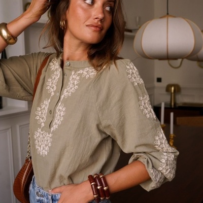 Blusa verde-acinzentada bordada com padrão floral branco em ambiente interior.