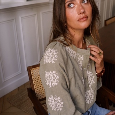 Mulher sentada com blusa verde-oliva bordada e acessórios dourados