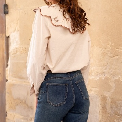 Camisa branca com gola bordada e calças de ganga azul escuro junto a parede de pedra