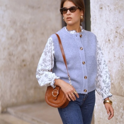 Mulher com camisola de malha azul e blusa branca com padrão floral, calças de ganga, óculos de sol, acessórios dourados e mala castanha ao ombro junto a parede de pedra clara.