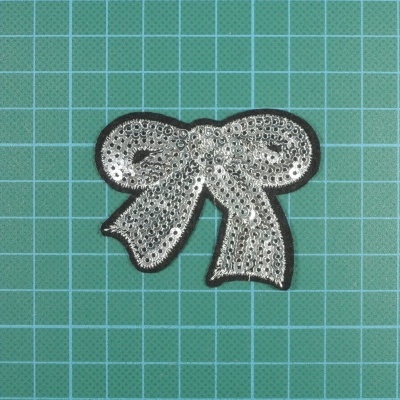 Aplique em forma de laço com lantejoulas prateadas sobre fundo verde quadriculado