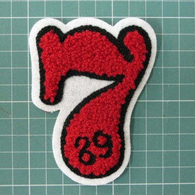 Emblema bordado em forma de número 7 vermelho com 89 preto, sobre fundo quadriculado verde