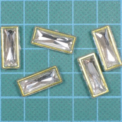 Cinco enfeites retangulares com pedras transparentes e moldura dourada sobre superfície azul quadriculada