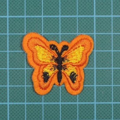 Pala bordada em forma de borboleta em laranja e amarelo sobre fundo verde xadrez.