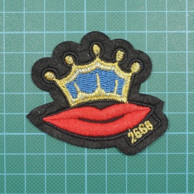 Emblema bordado com coroa dourada e lua azul sobre lábios vermelhos em fundo verde quadriculado