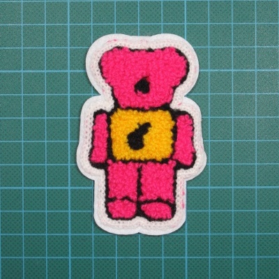 Adesivo bordado urso rosa com detalhe amarelo em fundo branco sobre superfície verde quadriculada