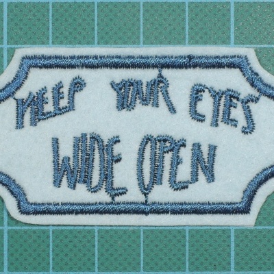 Etiqueta bordada azul com texto Keep Your Eyes Wide Open sobre fundo verde com grelha branca