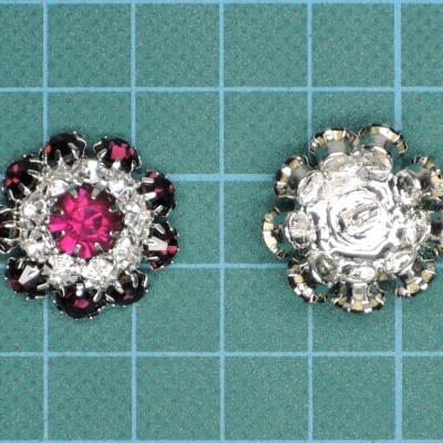 Dois botões decorativos redondos metal prateado com pedras vermelhas e rosas em superfície quadriculada azul