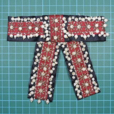 Laço étnico decorativo vermelho, preto e dourado com pérolas num fundo quadriculado verde.