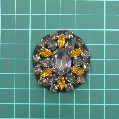 Adereço oval com pedras cinza, amarelas e transparentes numa superfície quadriculada