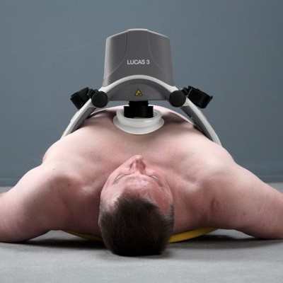 Homem deitado no chão com dispositivo LUCAS 3 sobre o peito, fundo cinzento.