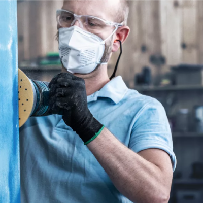 Homem a usar lixadora elétrica num objeto azul em oficina, com equipamentos de proteção.