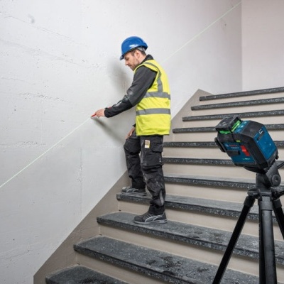 Homem com colete refletor medindo parede em escadaria com tripé e laser