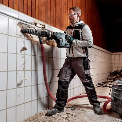 Homem a usar martelo demolidor elétrico num espaço com azulejos brancos e equipamento Metabo.