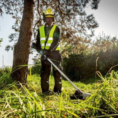 Pessoa com colete refletor e capacete a cortar erva ao ar livre