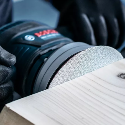 Lixadora orbital preta, vermelha e azul a lixar madeira clara com luvas pretas