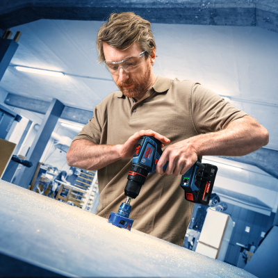 Homem usando furadeira BOSCH azul e preta numa superfície plana cinzenta em ambiente industrial