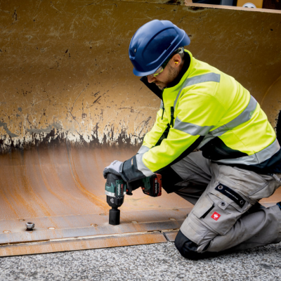 Trabalhador com capacete azul e colete amarelo usando parafusadeira numa superfície junto a estrutura metálica