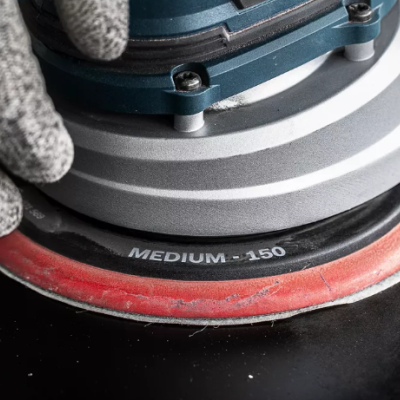 Lixadora elétrica circular com disco vermelho e preto, texto 'MEDIUM - 150', mão com luva cinza segurando o aparelho sobre superfície escura.