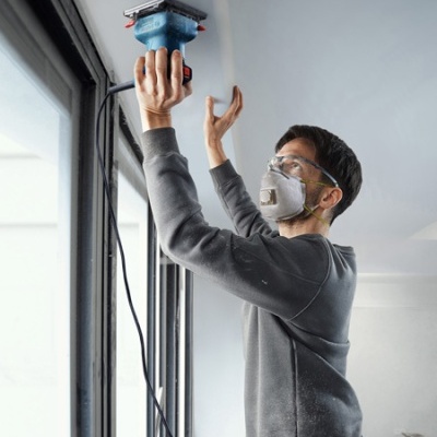 Pessoa a lixar o teto com lixadora azul, usando máscara e óculos