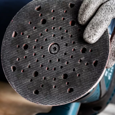 Disco de lixagem preto redondo com vários furos segurado por mão com luva cinza