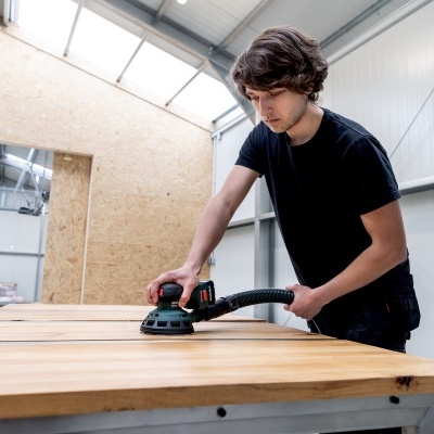 Homem a lixar madeira com lixadora elétrica numa oficina.